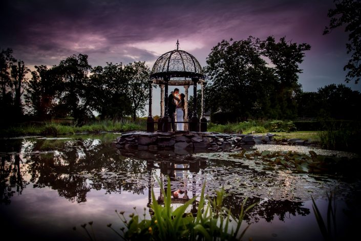 bride and groom standing under a glass archway by a lake